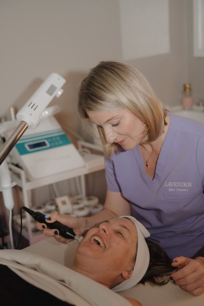 Esthetician in lavender scrubs giving a facial to a client wearing a headband