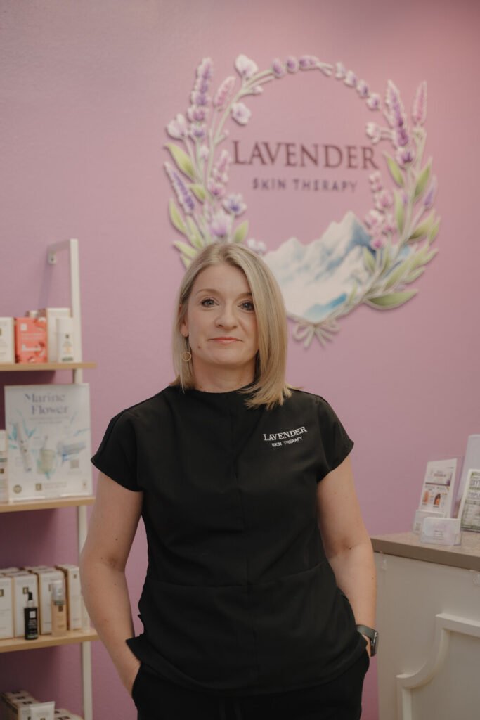 Joanna Bismark-Pettit, founder of Lavender Skin Therapy, standing in front of the studio logo wall